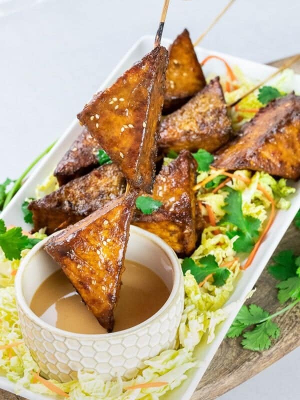 baked thai tofu satay skewers dipped in peanut satay sauce on a white plate with wooden board