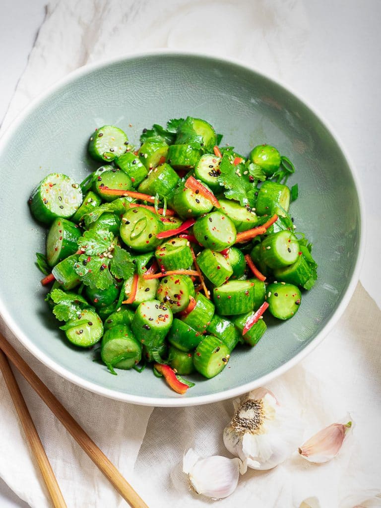Spicy Asian cucumber salad with herbs and red pepper in a blue bowl next to garlic and chopsticks