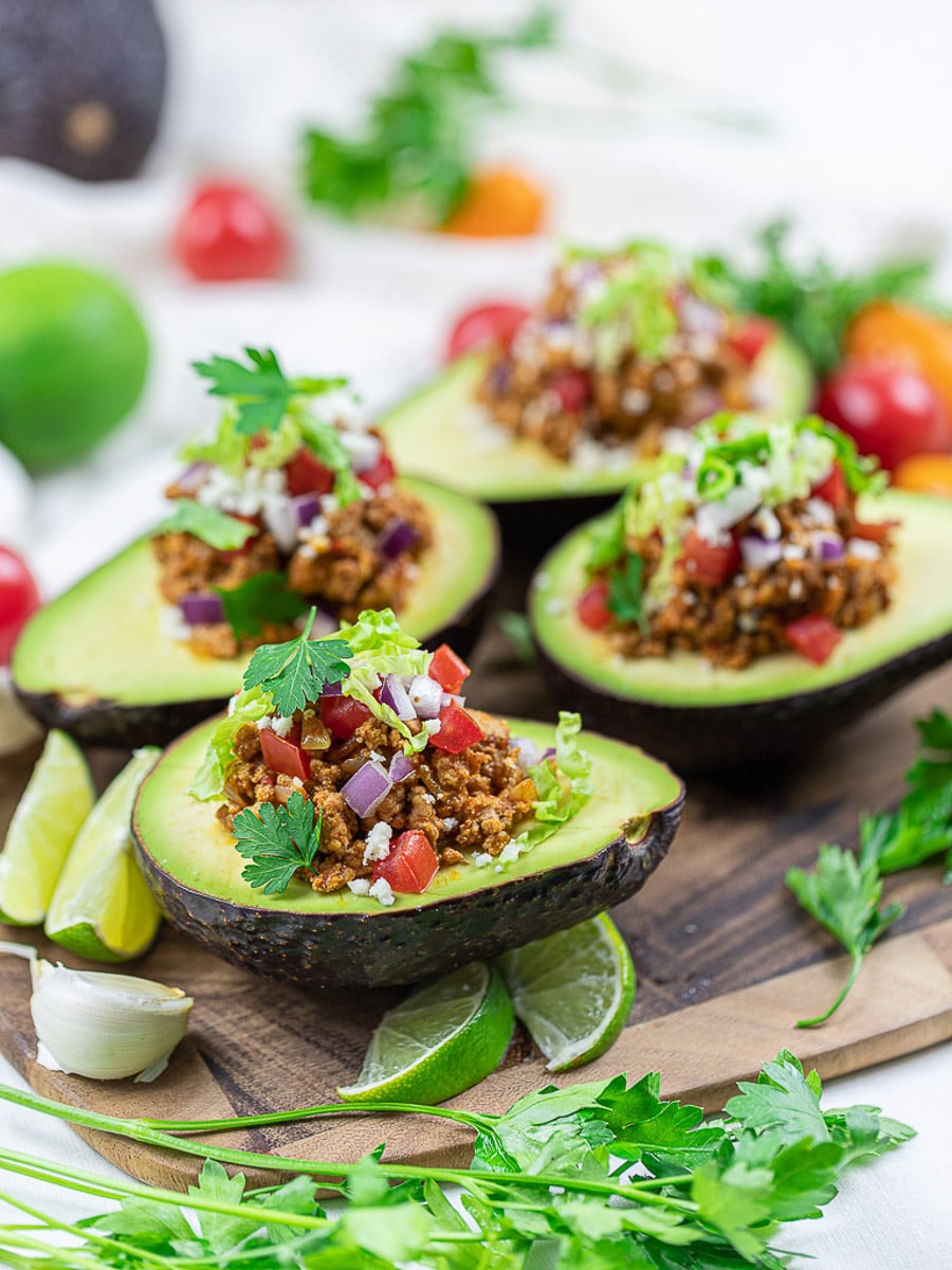 Keto taco avocado boats with lime, taco meat, tomatoes, and cilantro on a wooden board.