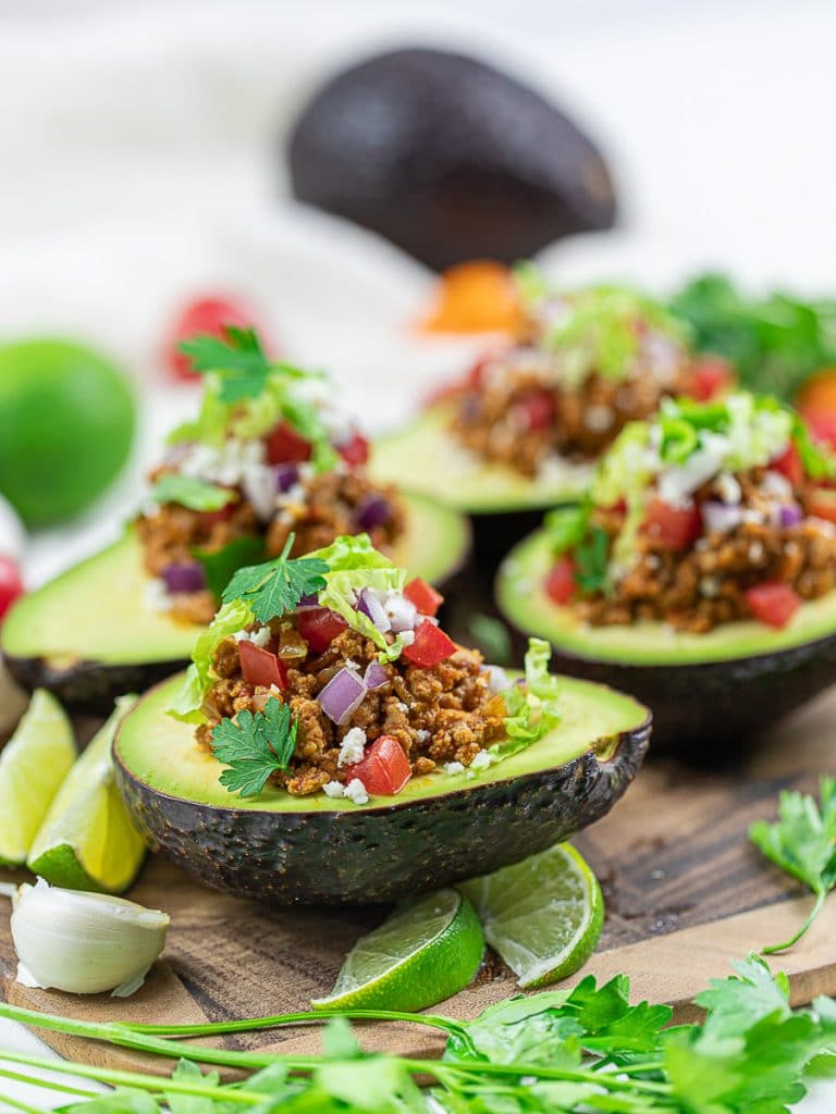 Keto Taco Stuffed Avocado boats on a wooden cutting board with lime, taco meat, tomatoes, and cilantro