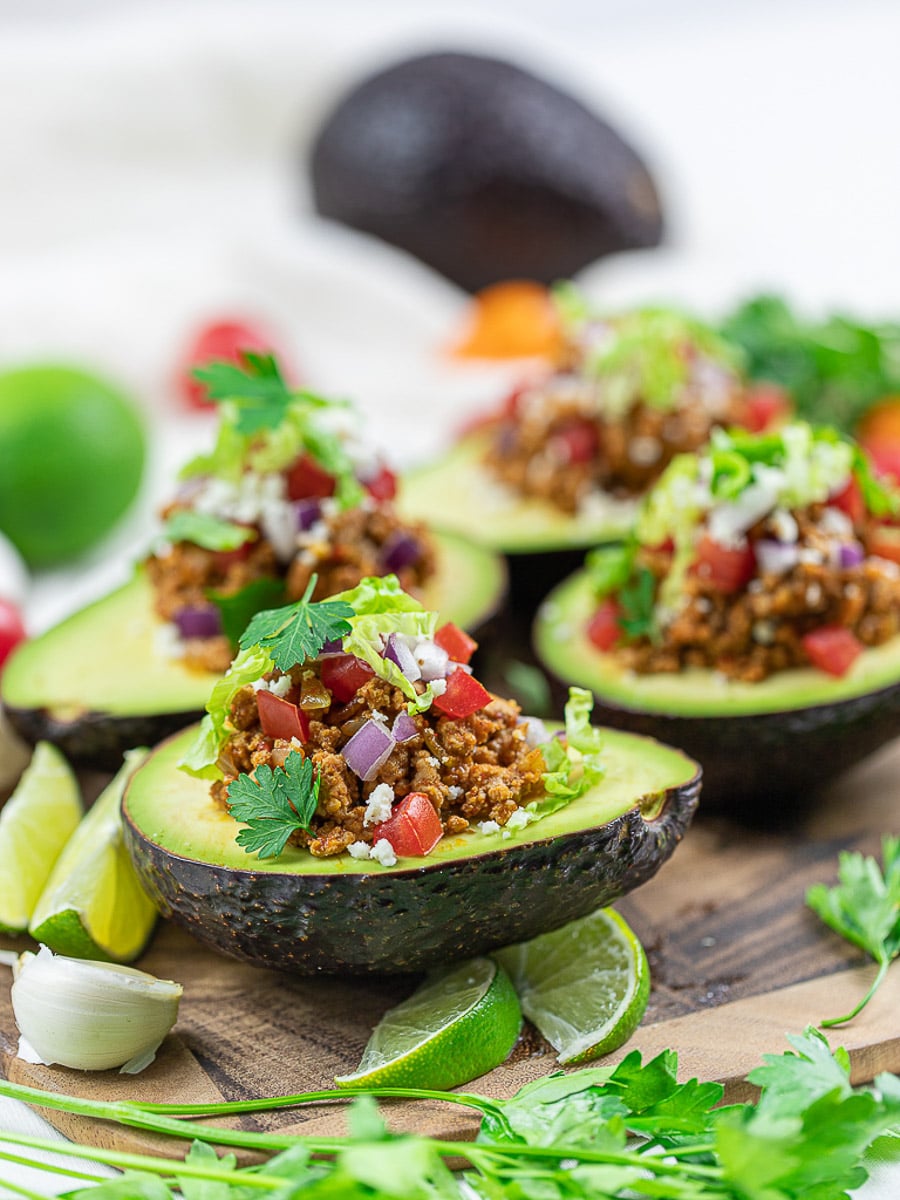 Keto taco avocado boats on a wooden cutting board with lime, taco meat, tomatoes, and cilantro.