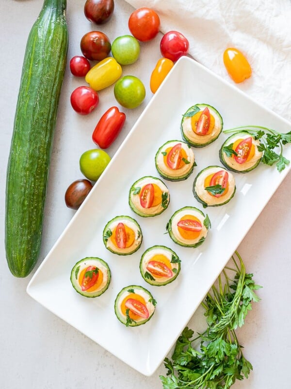 vegan cucumber hummus bites with red and orange cherry tomatoes on a white plate