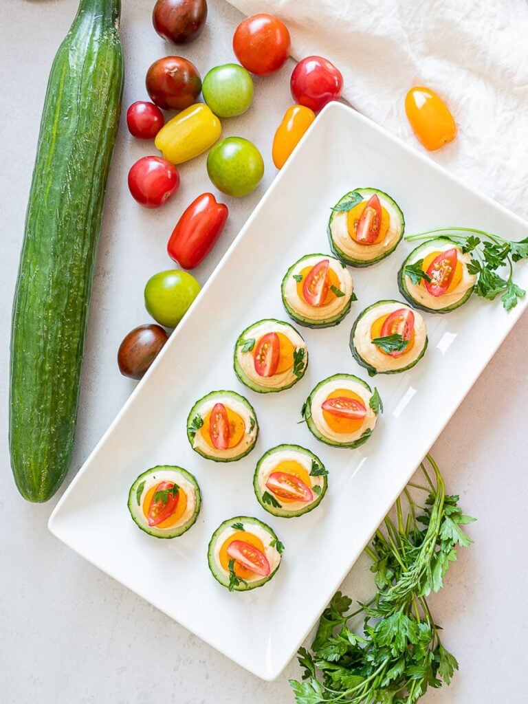 vegan cucumber hummus bites with red and orange cherry tomatoes on a white plate