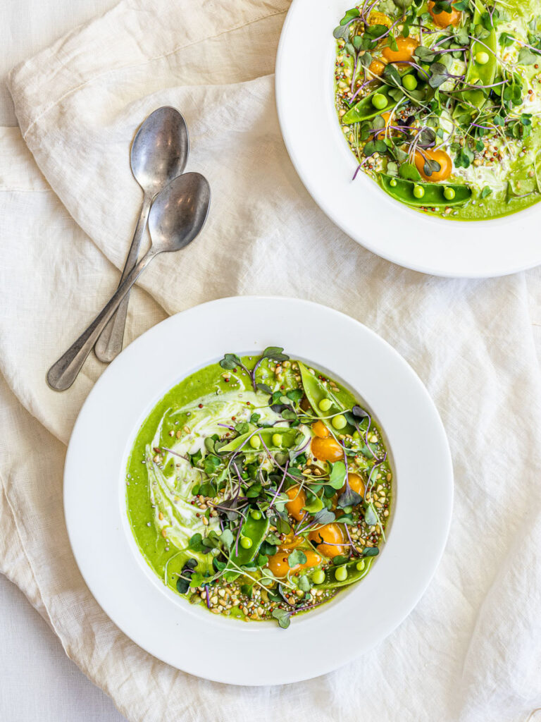 Two bowls of chilled vegan cucumber avocado soup with peas, tomatoes, and microgreens in white bowls.
