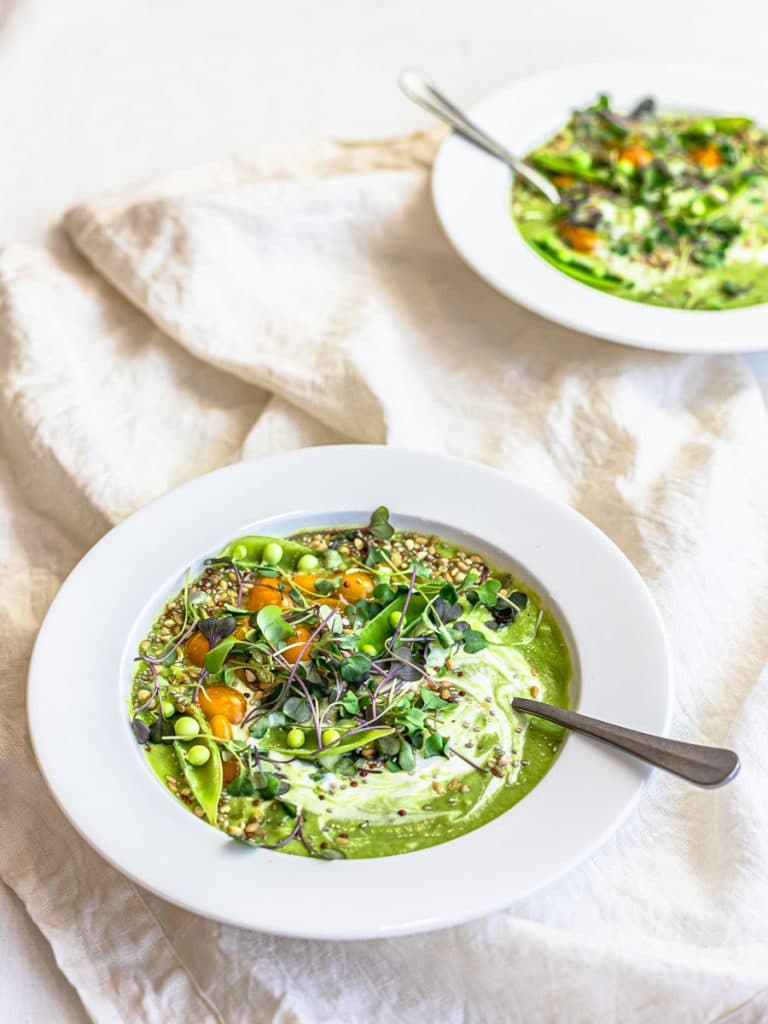 Two bowls of chilled vegan cucumber avocado soup with peas, tomatoes, and microgreens in white bowls.