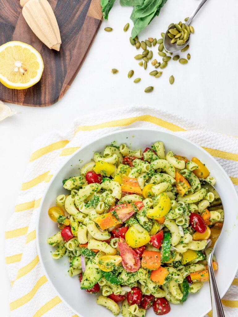 cold vegan pesto pasta salad with tomatoes and vegetables in a white bowl with lemon