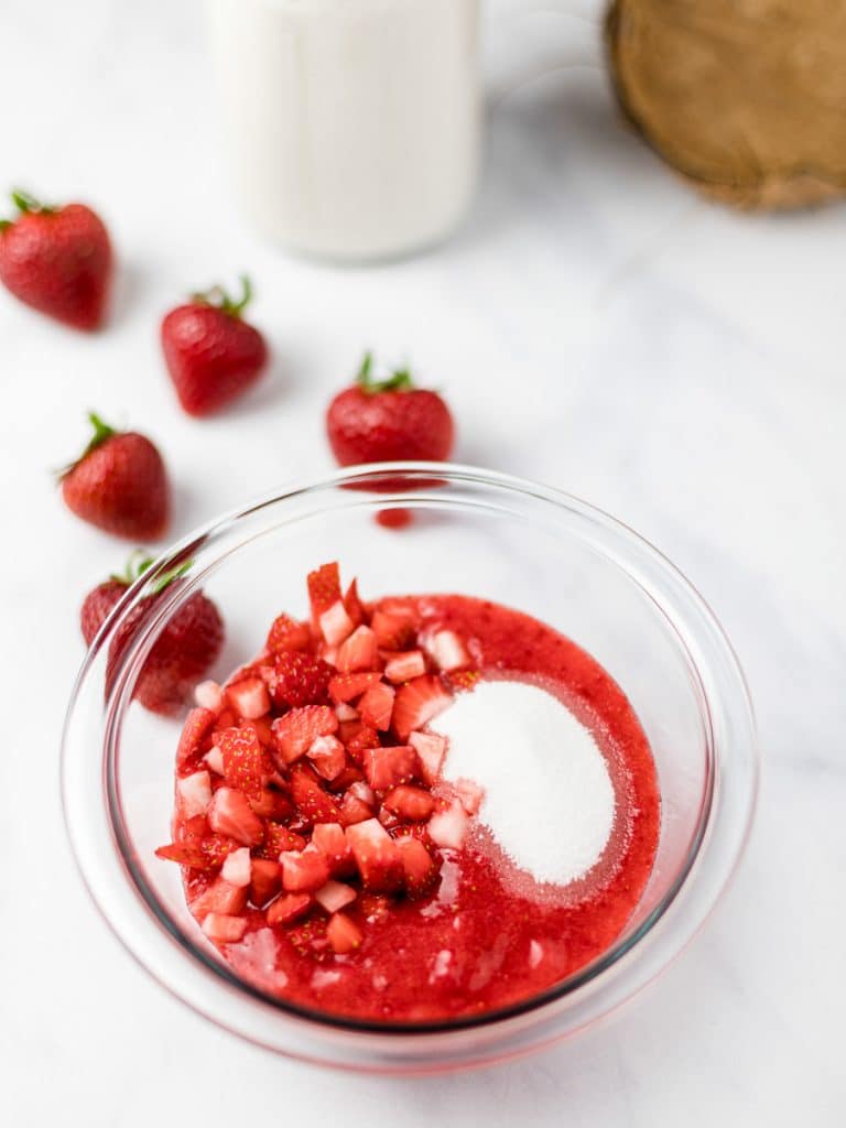 strawberry pieces, strawberry puree, and sugar in a glass bowl surrounded by strawberries