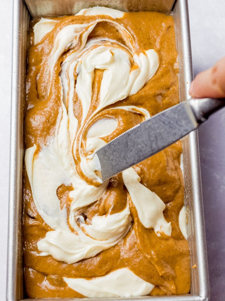 cream cheese being swirled into pumpkin bread batter in a loaf pan