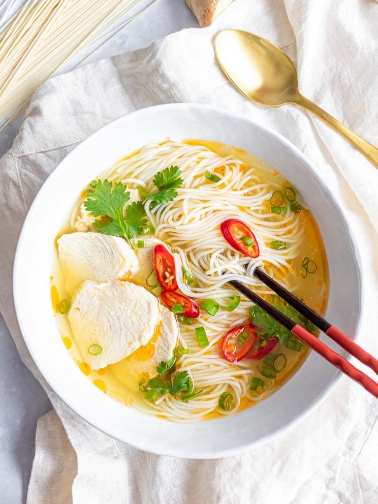 Asian chicken noodle soup with red peppers and herbs in a white bowl with chopsticks and a spoon