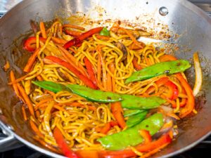 lo mein noodles with snow peas, red peppers, and carrots cooking in a wok