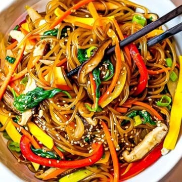 japchae, Korean glass noodles with red peppers, carrots, and shiitake mushrooms in a white bowl with chopsticks