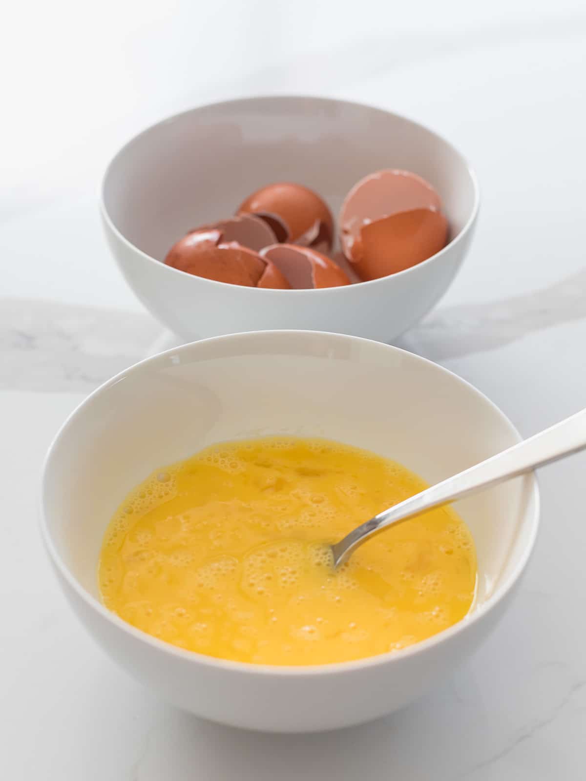 bowl of beaten eggs with a silver fork next to a bowl filled with egg shells