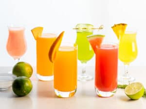different fruit flavors of aguas frescas including watermelon, cucumber, pineapple, mango, and cantaloupe next to limes