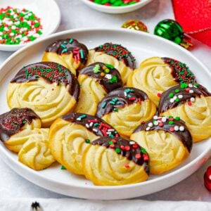 Christmas butter cookies dipped in chocolate with Christmas sprinkles on a white plate surrounded by decorations