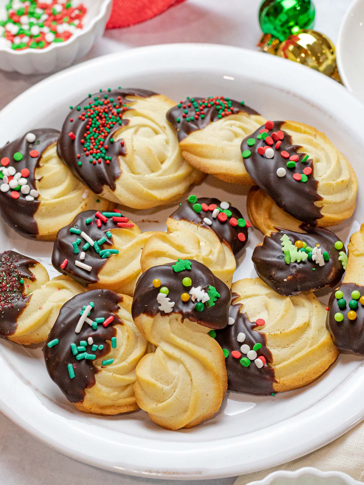 Christmas butter cookies dipped in dark chocolate with red and green sprinkles on a white plate surrounded by ornaments and more sprinkles