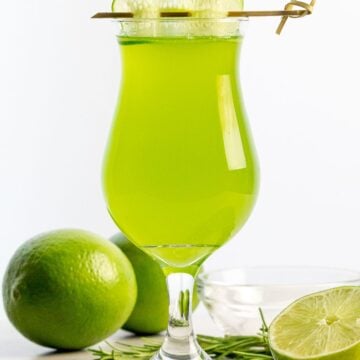 cucumber agua fresca with a cucumber garnish next to limes and herbs