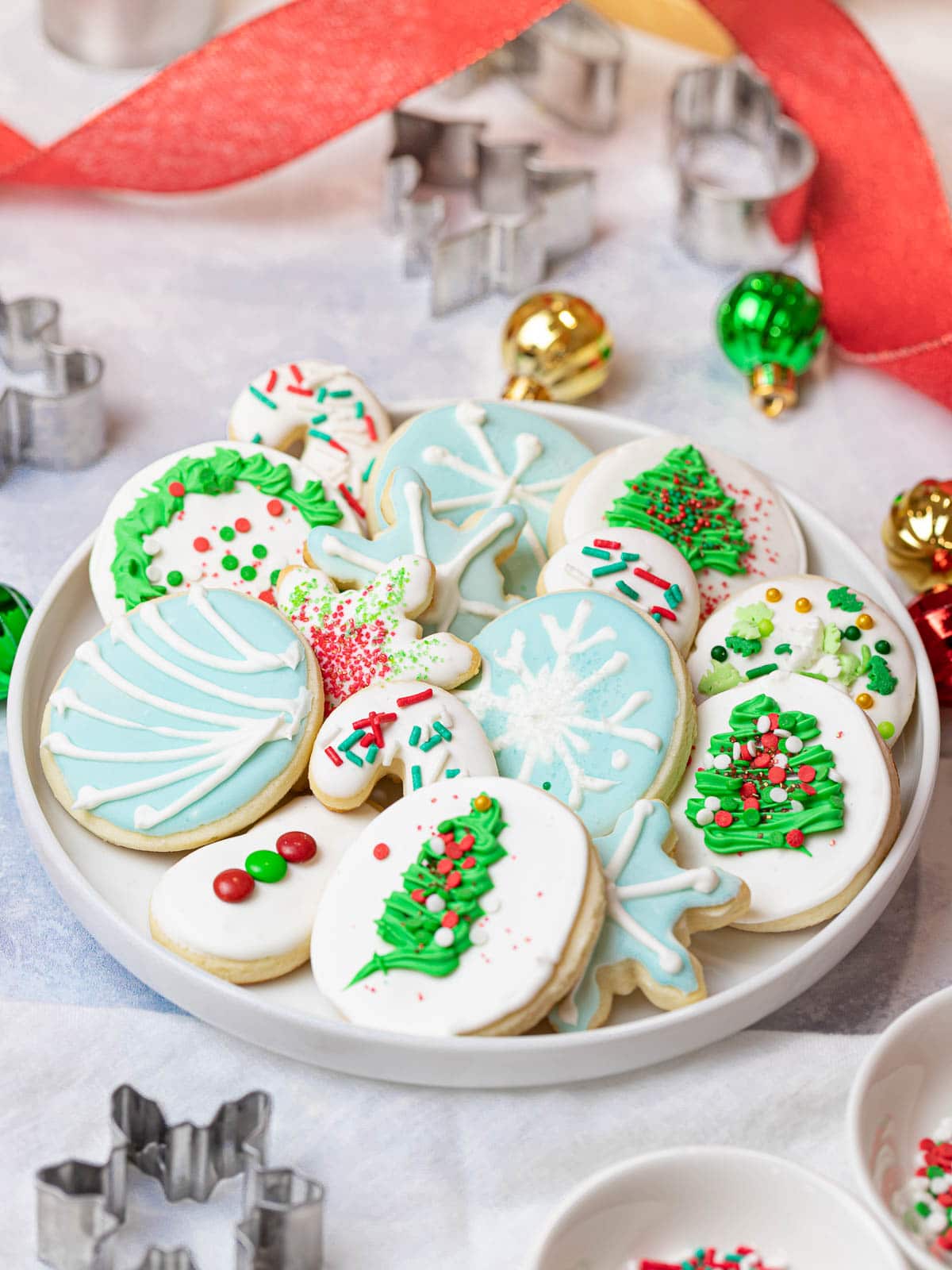 easy Christmas sugar cookies decorated with sprinkles and icing placed on a plate next to ornaments, cookie cutters, and red ribbon