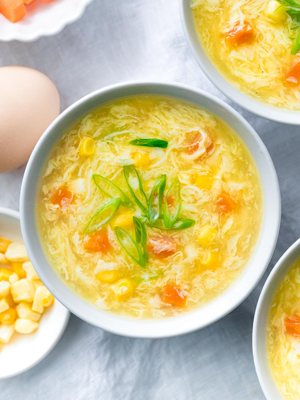 egg drop soup with corn and carrots in a bowl next to corn kernels and an egg