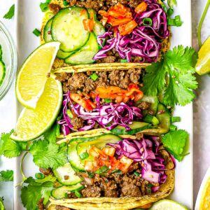 Korean beef tacos with cabbage slaw, kimchi, cucumbers, and lime on a white plate