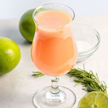 glass of peach agua fresca next to limes and herbs