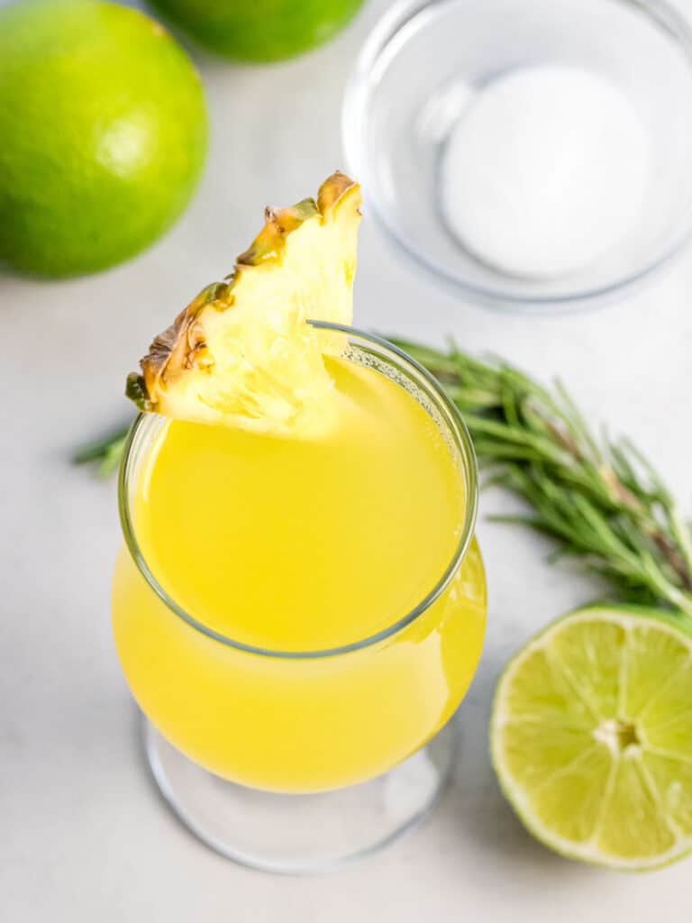 glass of pineapple agua fresca next to limes, herb, and sugar