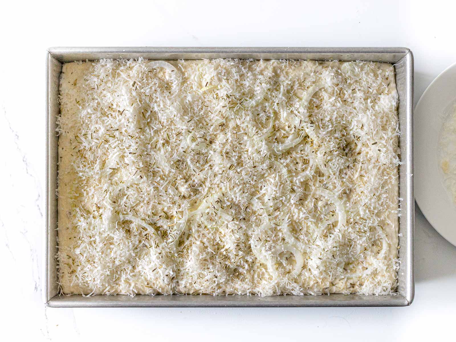 focaccia dough topped with parmesan cheese, onions, and rosemary in a baking dish