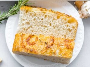 slice of rosemary parmesan focaccia bread on a white plate next to rosemary and garlic