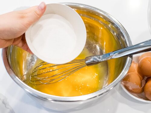 sugar being added to egg yolks in a metal bowl with a whisk