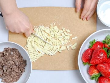 white chocolate being chopped by a knife on a cutting board next to chopped dark chocolate and strawberries in white bowls