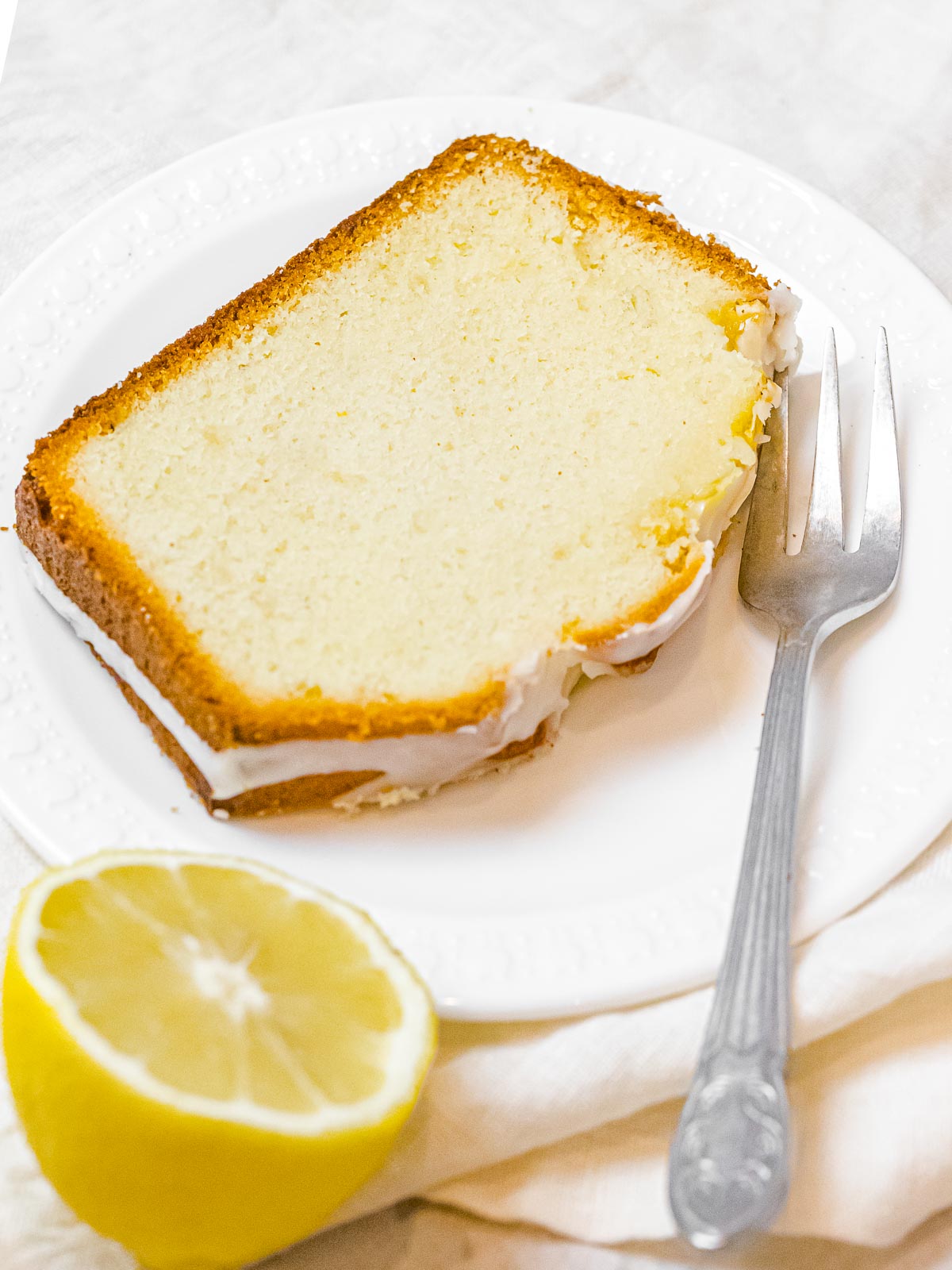 slice of lemon pound cake on a white plate next to a fork and half a lemon