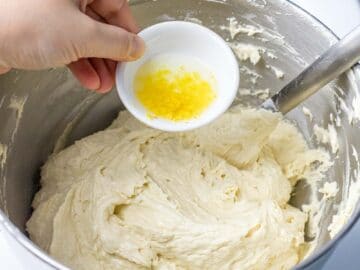 pound cake batter in a metal mixing bowl with lemon zest and lemon juice being added from a small white bowl