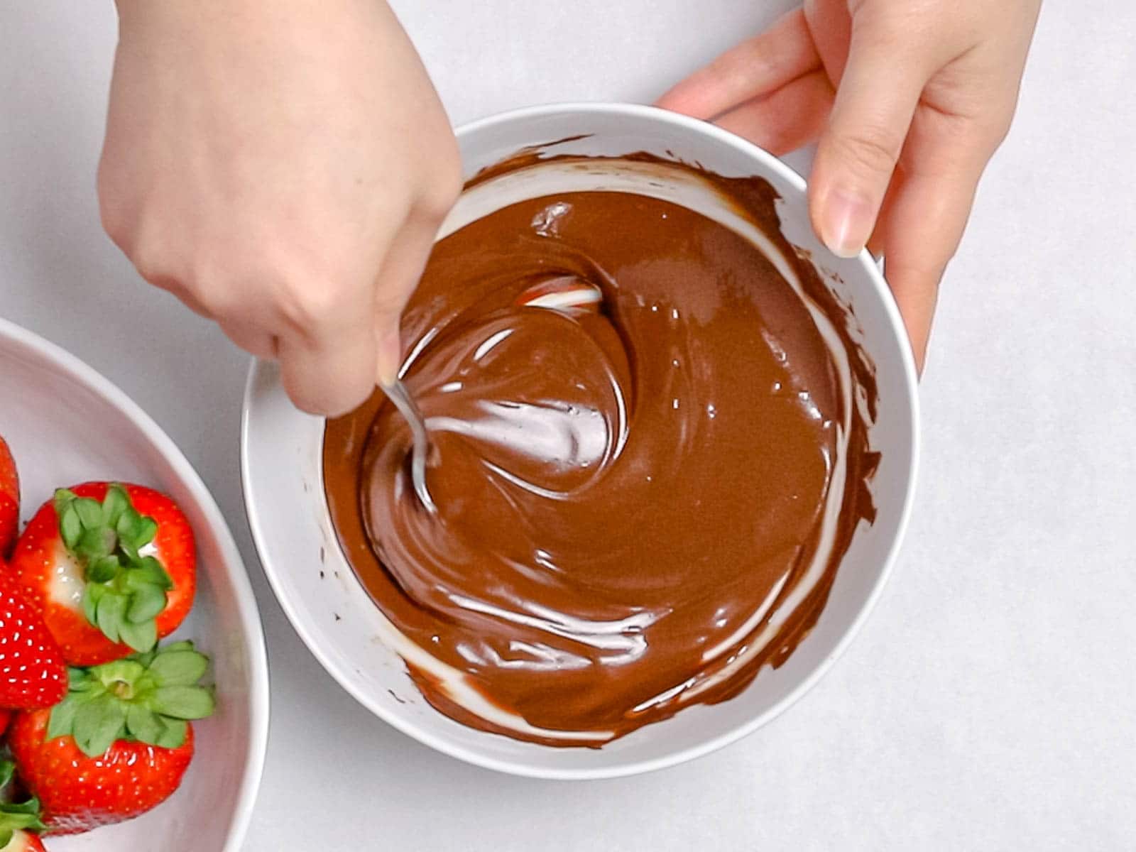 melted dark chocolate being stirred by a spoon in a white bowl next to a bowl of strawberries