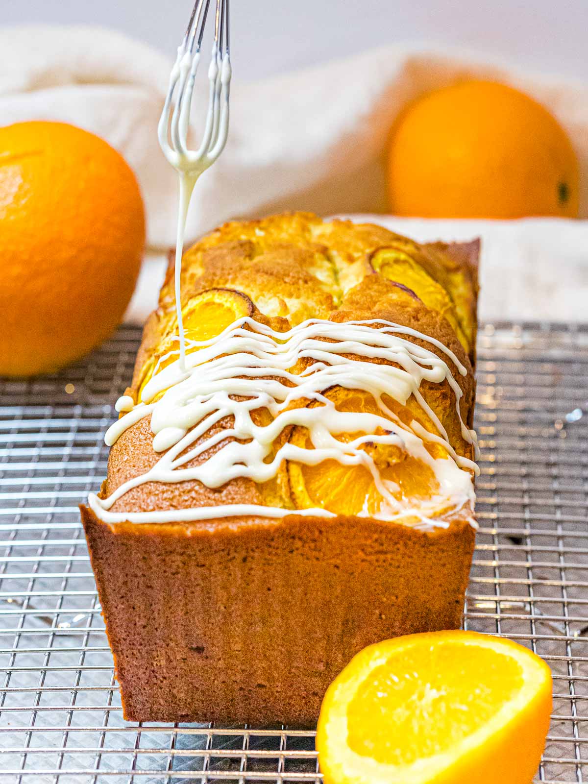 orange pound cake with glaze being drizzled on top with a whisk set on a wire rack next to oranges