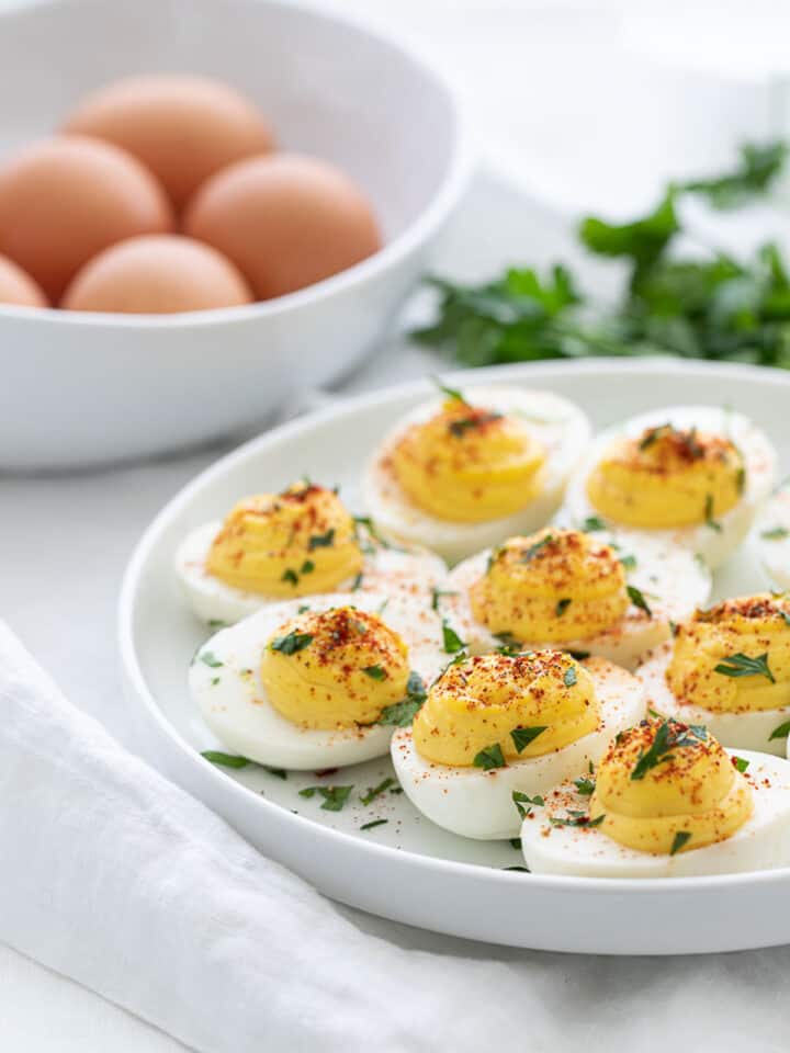 classic deviled eggs garnished with parsley and paprika on a white plate with a bowl full of brown shelled eggs in the background