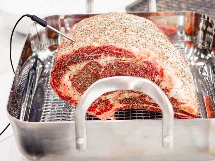 Prime rib roast with bone covered in salt and pepper rub in a roasting pan with a meat thermometer inserted before roasting.