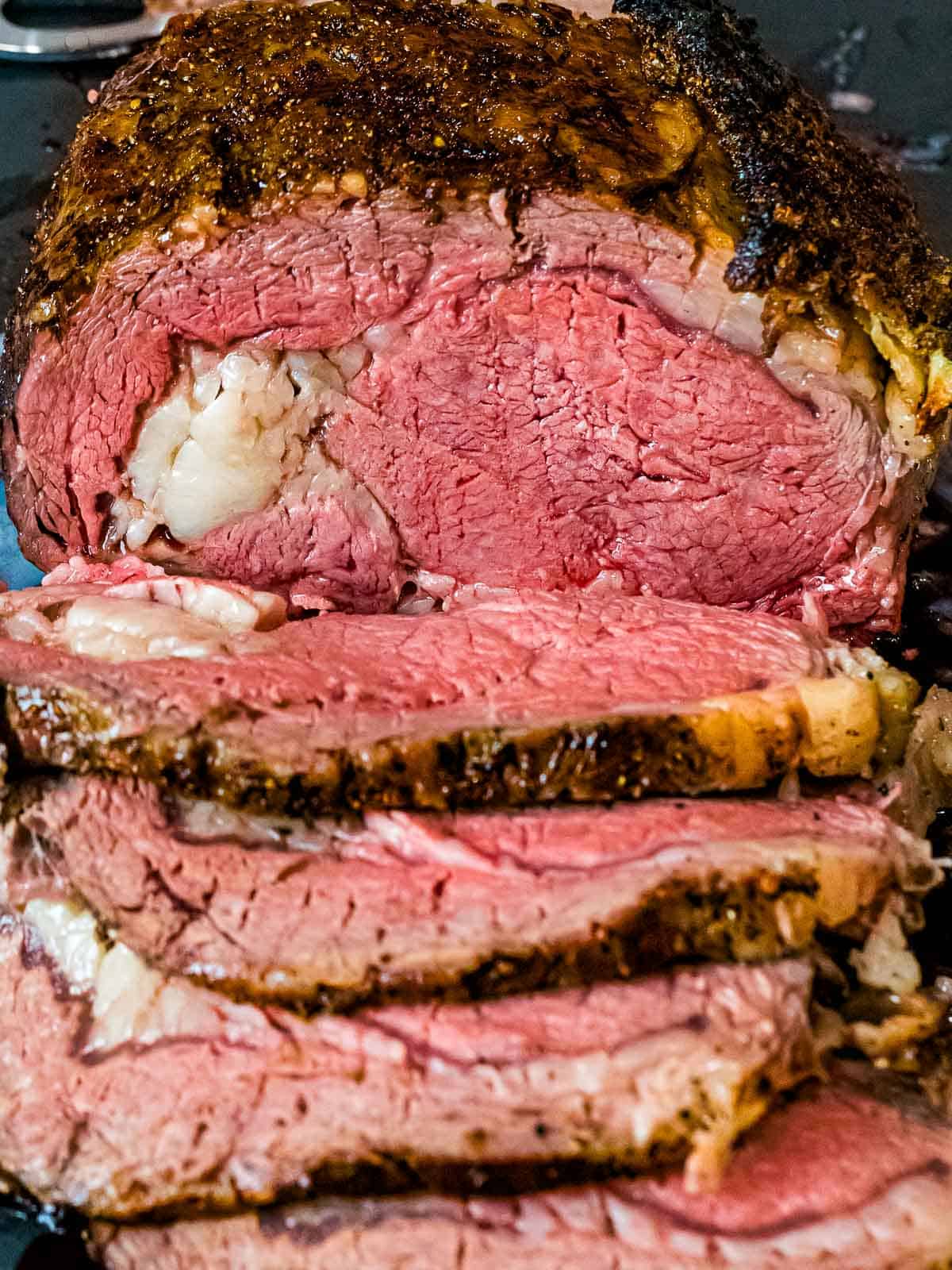Prime rib roast cooked to medium and sliced on a dark cutting board showing a crispy, crackly crust made from a salt and pepper rub.