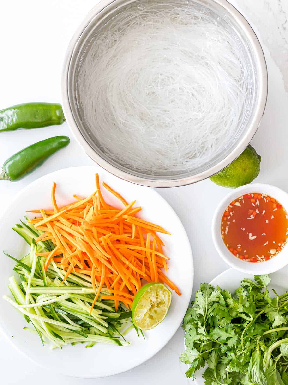 Vermicelli rice noodles in a stainless steel bowl next to carrots, cucumber, lime, vermicelli dressing, and herbs.