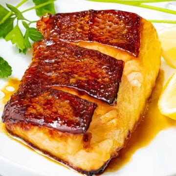 Chilean sea bass pan seared with golden brown crispy skin in an Asian marinade on a white plate with herbs and lemon.