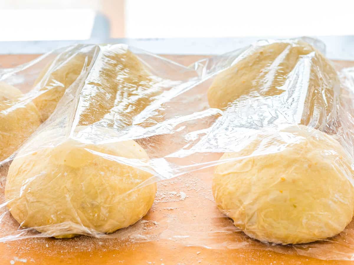 Dough formed into rounds on a wooden board dusted with flour and lightly covered with plastic wrap.