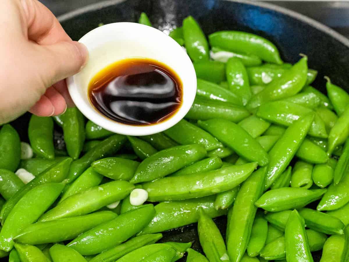 Soy sauce being added to sugar snap peas sauteed in a pan.