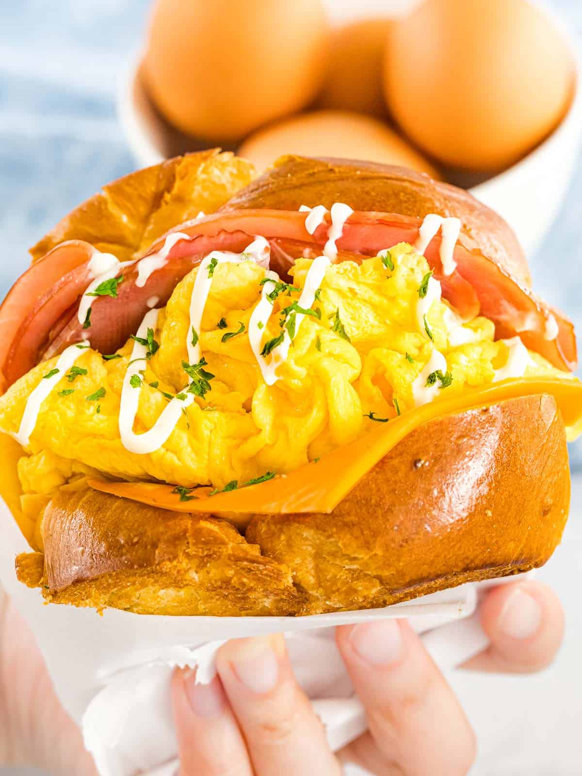 Hand holding a Korean egg drop sandwich with fluffy scrambled eggs, cheese, ham, and buttered toast with mayo drizzle next to bowl of eggs.