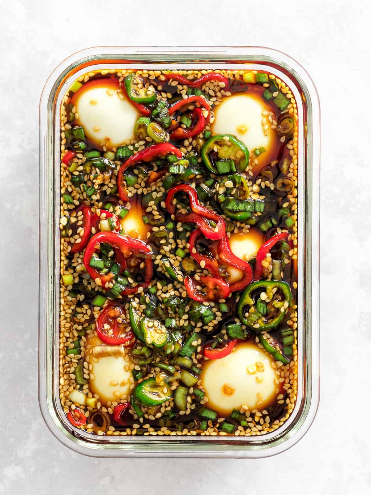 Mayak eggs in a soy marinade with red and green chili peppers and sesame seeds in a rectangular glass container.