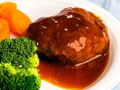 Juicy Japanese hamburger steak (hambāgu ハンバーグ) covered in sauce on a white plate next to carrots and broccoli.