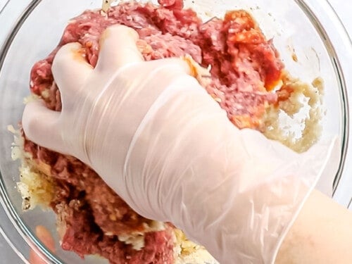 Hamburger steak ingredients in a glass bowl being mixed together with a gloved hand.