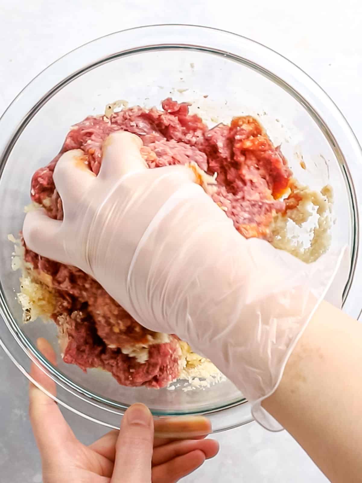 Hamburger steak ingredients in a glass bowl being mixed together with a gloved hand.