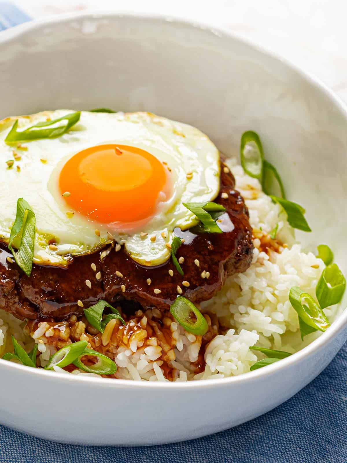 Loco moco in a white bowl filled with white rice, beef burger, brown gravy, and a fried egg that is garnished with green onions.