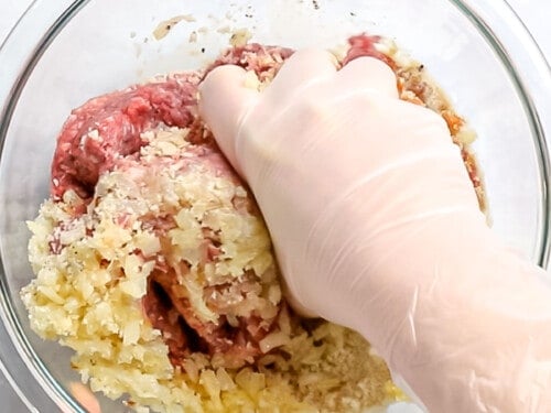 Loco moco patty being mixed with ground beef, onions, bread crumbs, and seasonings by a gloved hand.