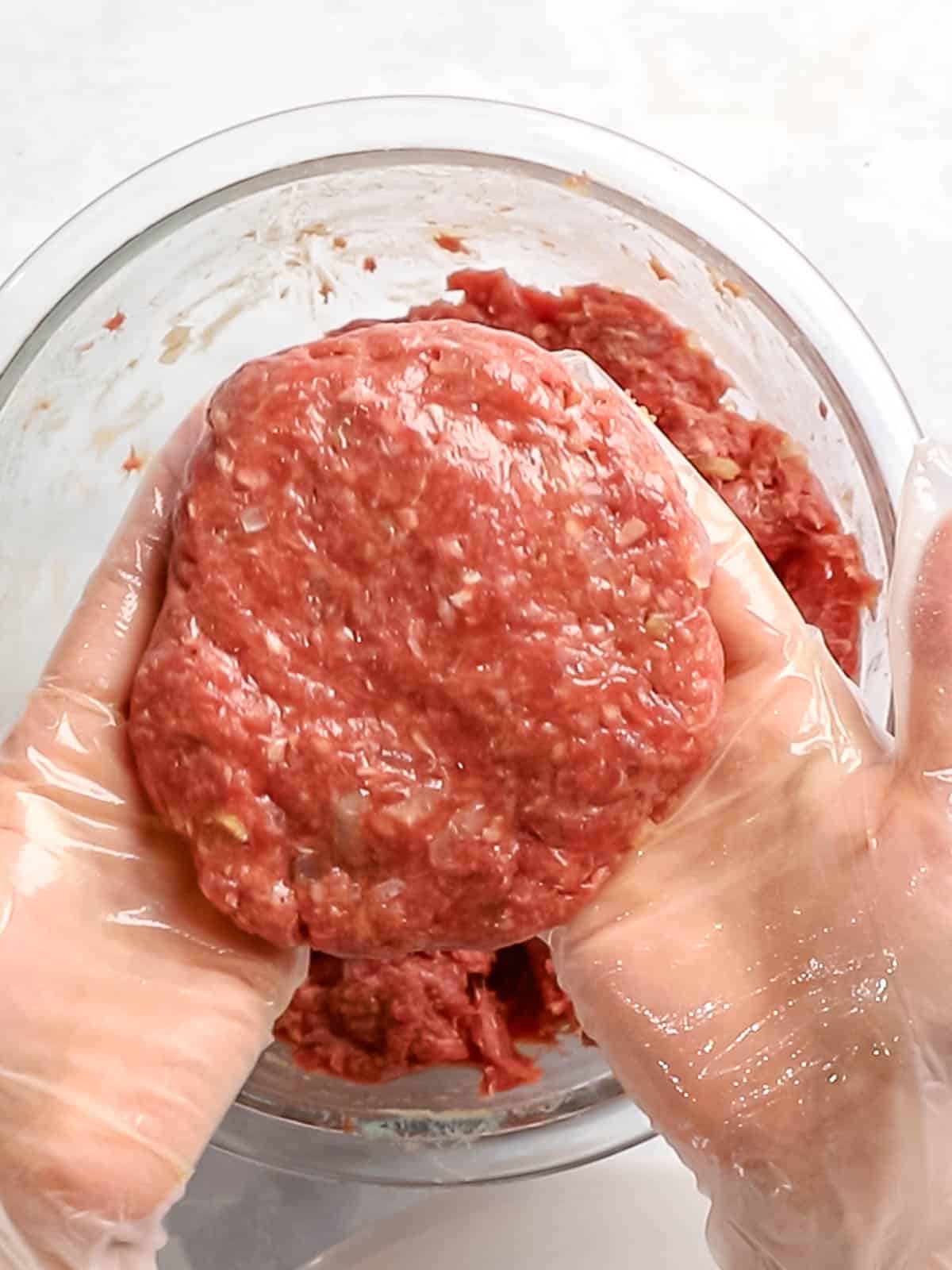 Ground beef formed into a burger patty that is held with gloved hands.