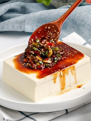 Soy sauce and green onions being poured over silken tofu with a wooden spoon.