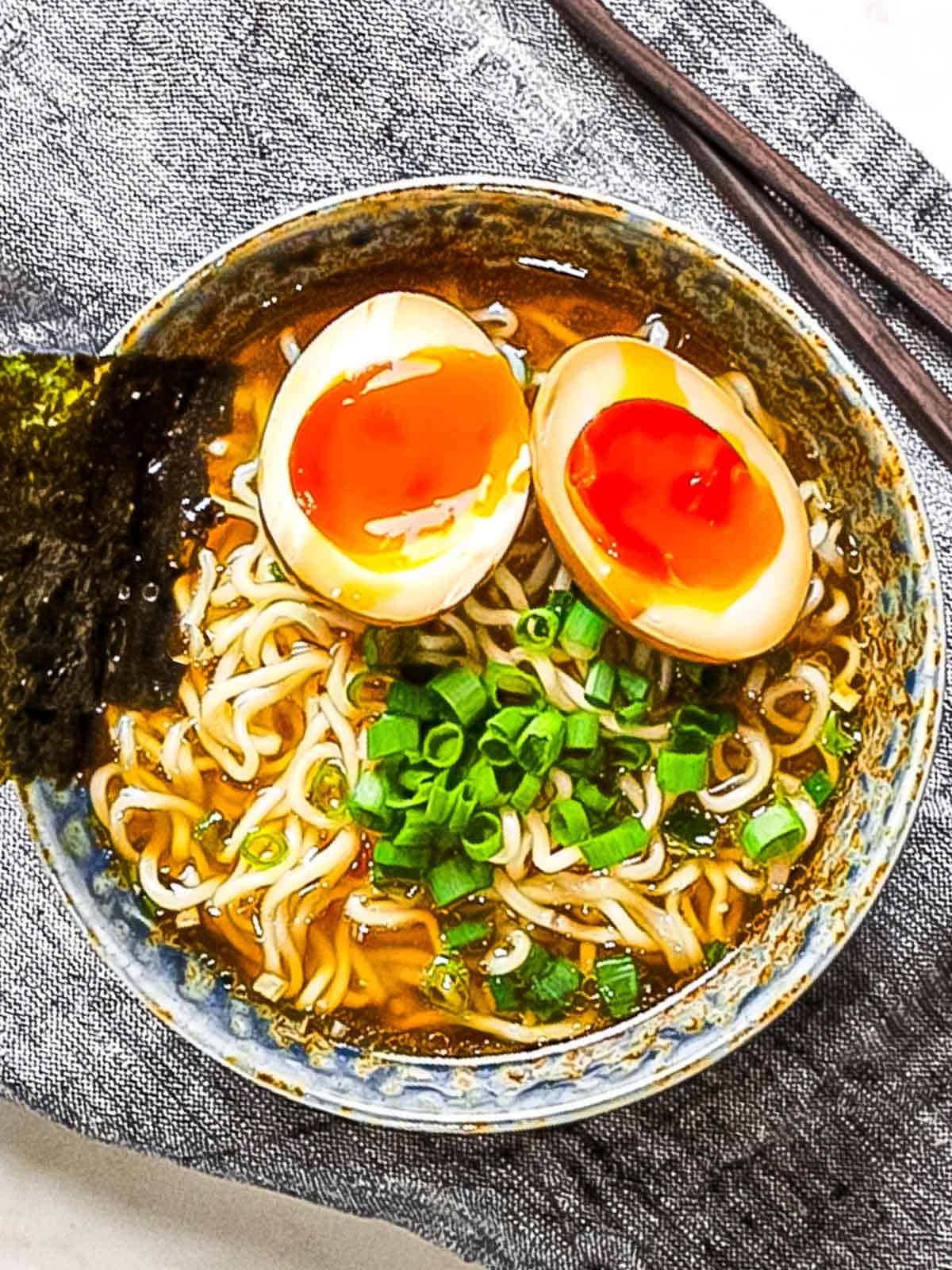 Ramen eggs (ajitama) marinated in soy sauce cut in half and placed on top of a bowl of ramen noodles garnished with green onions and nori.